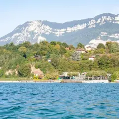 Vue sur le lac du Bourget, avec des maisons sur la colline verdoyante. Un chien court sur la plage, profitant de la journée.
