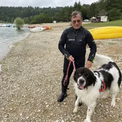 Homme en combinaison de plongée avec son chien noir et blanc sur la plage du lac de Vouglans. Chien autorisé, prêt pour une baignade.