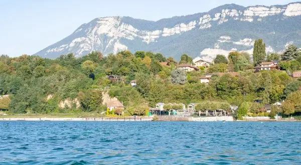 Vue sur le lac du Bourget, avec des maisons sur la colline verdoyante. Un chien court sur la plage, profitant de la journée.