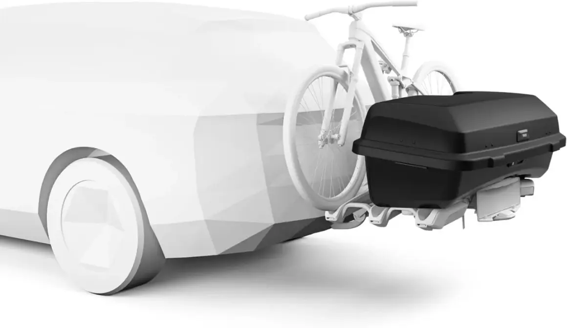 Un coffre sur attelage noir est fixé à l'arrière d'une voiture blanche stylisée, avec un vélo blanc monté juste devant.