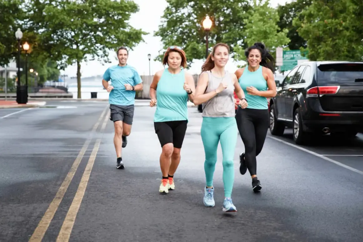 Un groupe de quatre personnes, deux hommes et deux femmes, commence la course à pied sur une route mouillée, entourés d'arbres et de lampadaires.