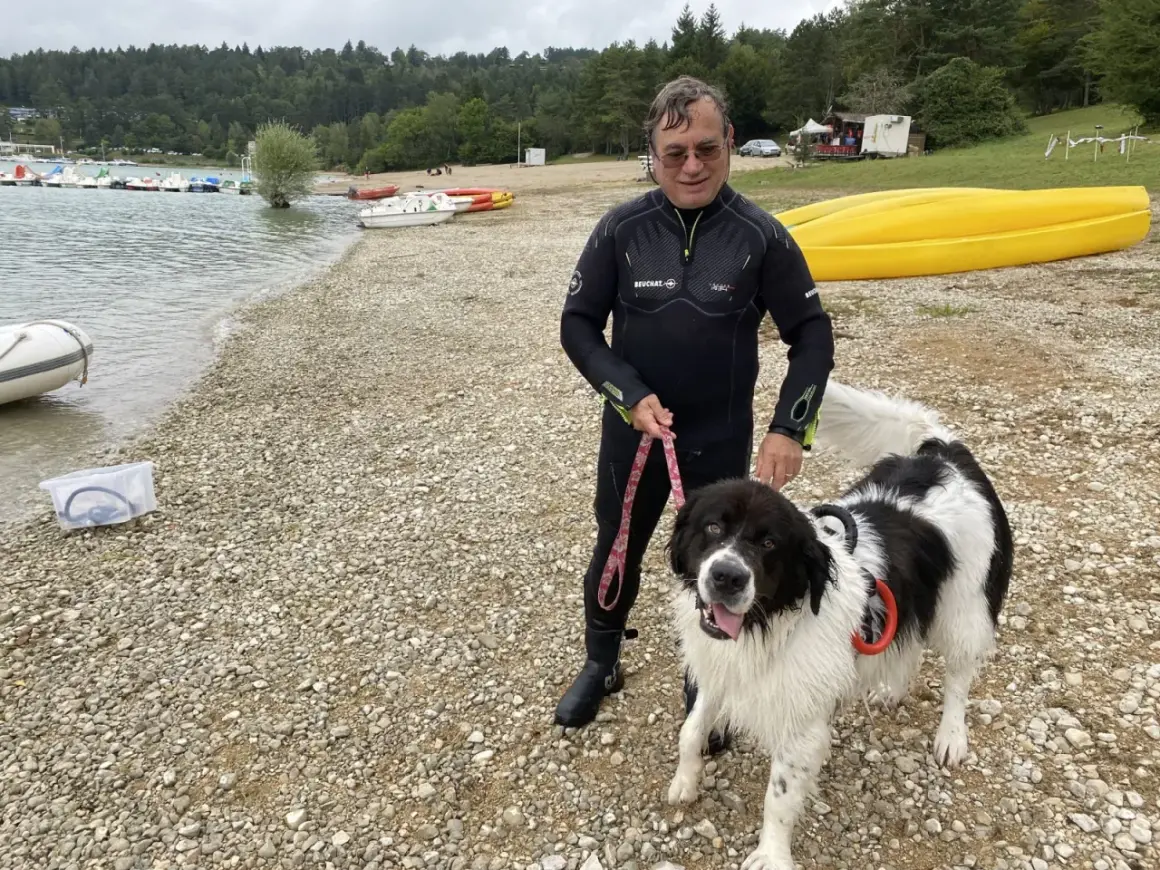 Homme en combinaison de plongée avec son chien noir et blanc sur la plage du lac de Vouglans. Chien autorisé, prêt pour une baignade.