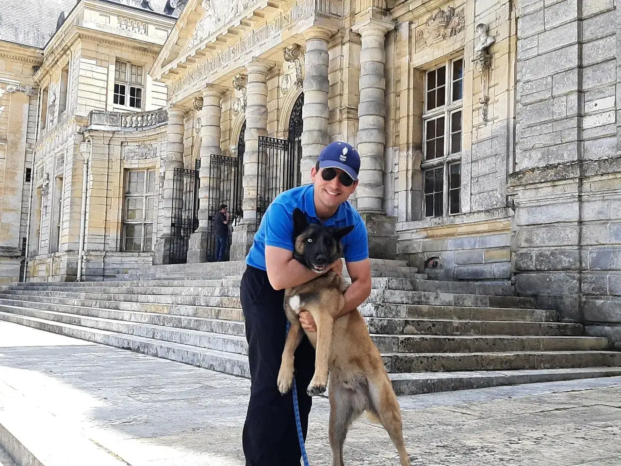 Un homme et son chien profitent d'une visite au château. Le chien est tenu dans les bras de l'homme devant un grand escalier.