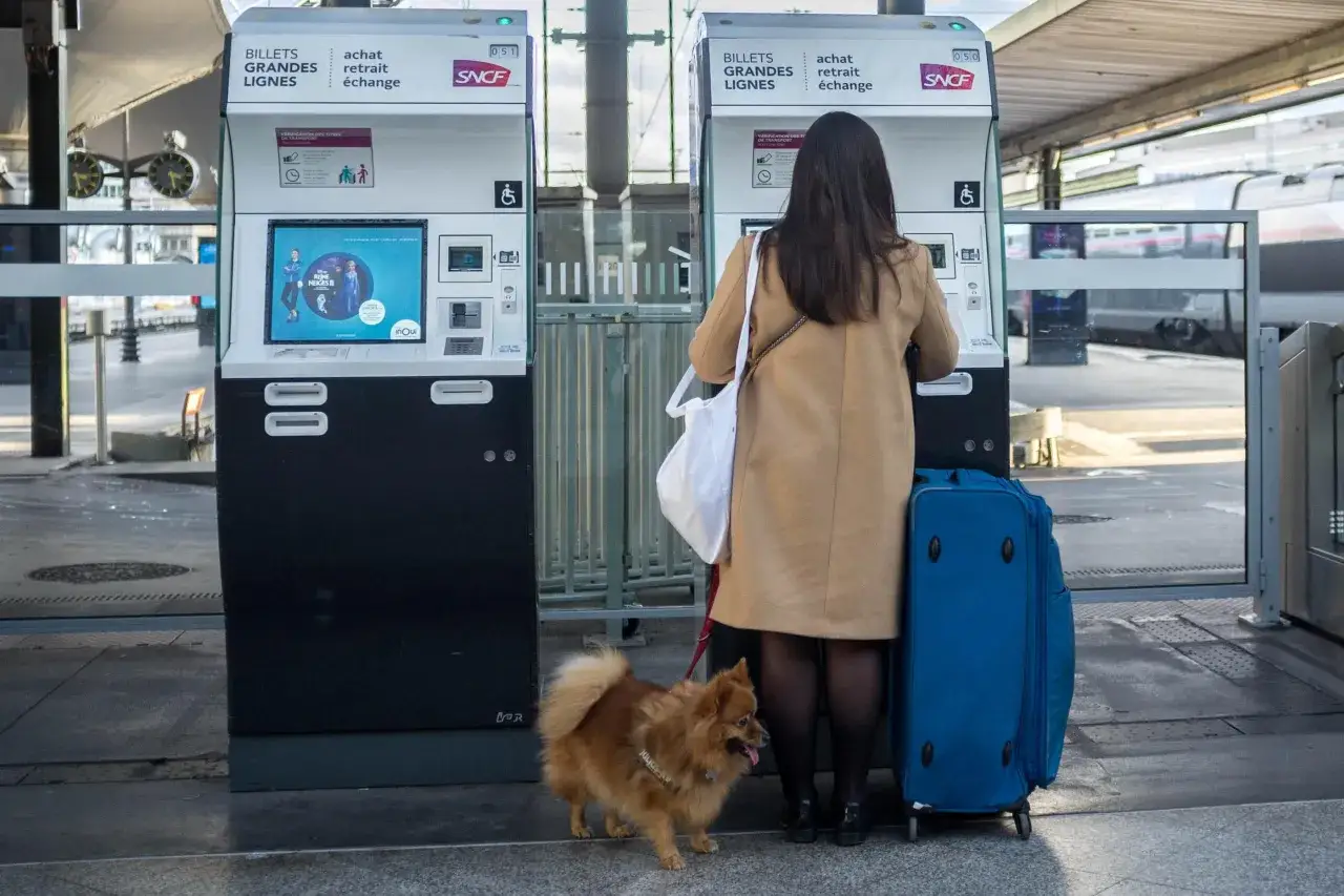 Une femme achète un billet à la gare SNCF, son chien à ses côtés. Espérons qu'elle n'ait pas d'amende chien SNCF pour son compagnon à quatre pattes.