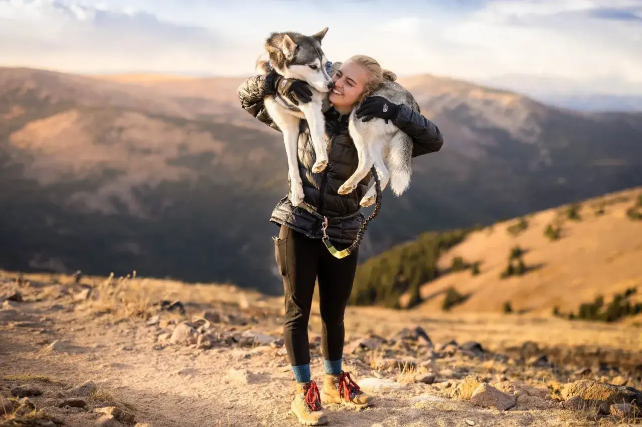 Une femme porte son chien dans la neige, sur une montagne. Le chien est un husky, et il semble heureux.