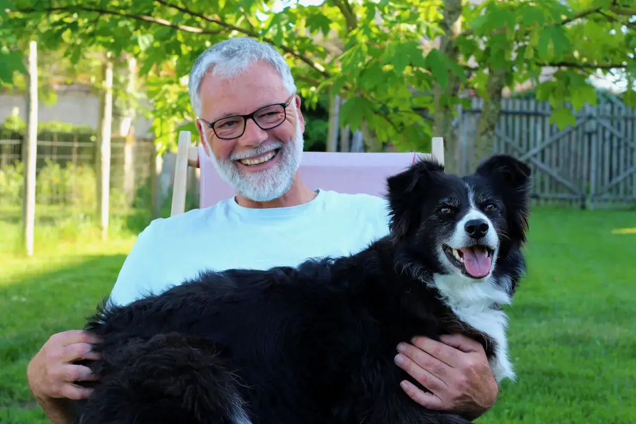 Un homme souriant, un éducateur canin comportementaliste, tient son chien noir et blanc dans ses bras, profitant du soleil dans un jardin.