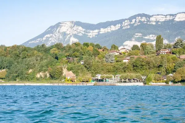 Vue sur le lac du Bourget, avec des maisons sur la colline verdoyante. Un chien court sur la plage, profitant de la journée.
