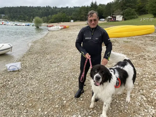 Homme en combinaison de plongée avec son chien noir et blanc sur la plage du lac de Vouglans. Chien autorisé, prêt pour une baignade.